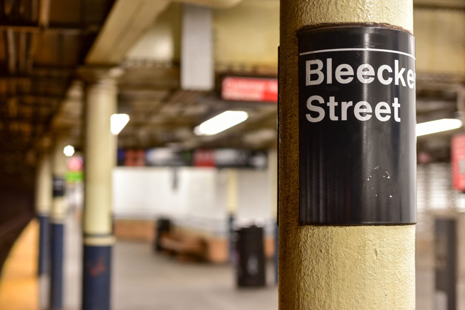 A bleecker street sign on a column in a subway station, with softly focused platform and benches in the background.