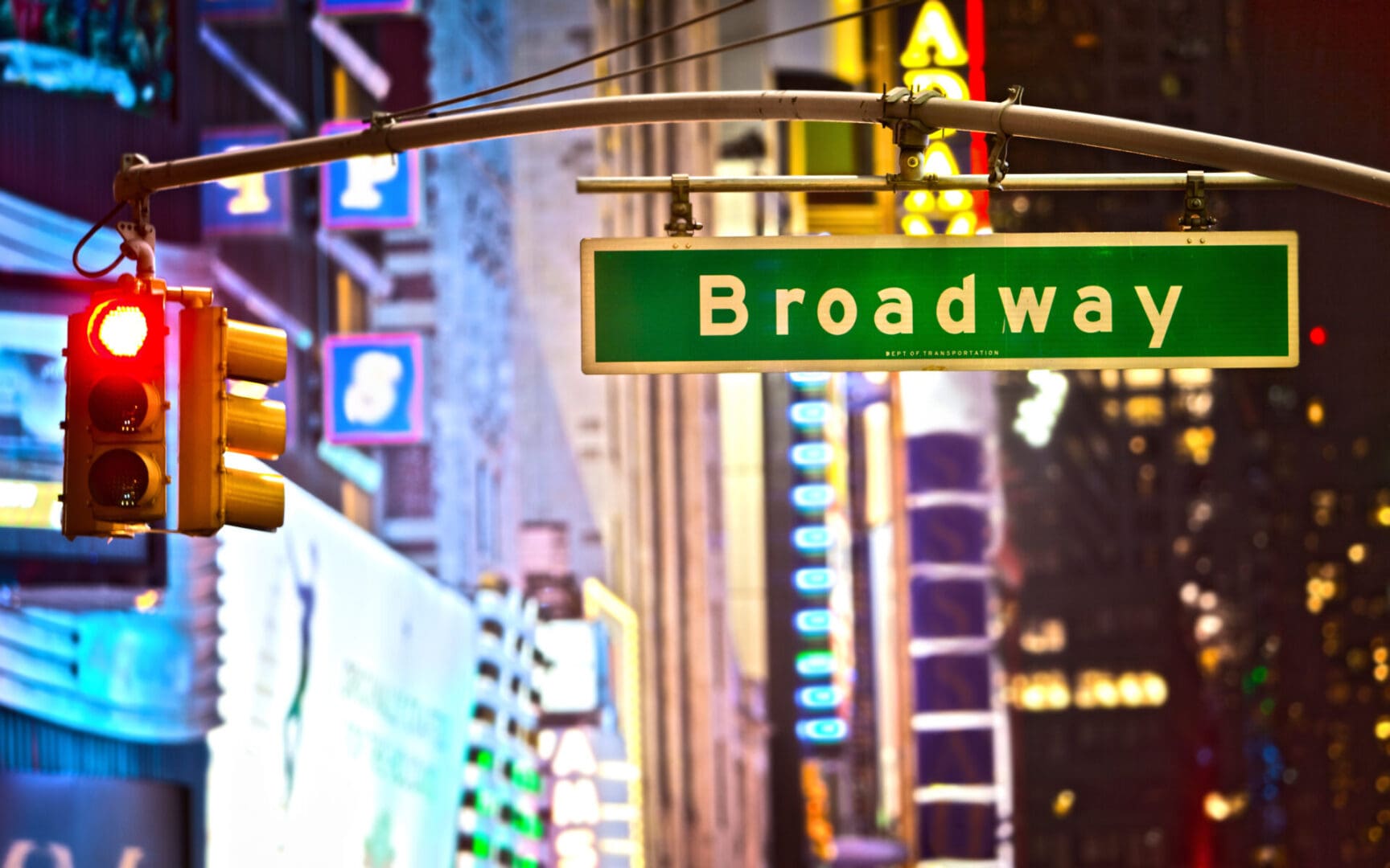 Illuminated "broadway" street sign and traffic light at night with blurred city lights in the background.