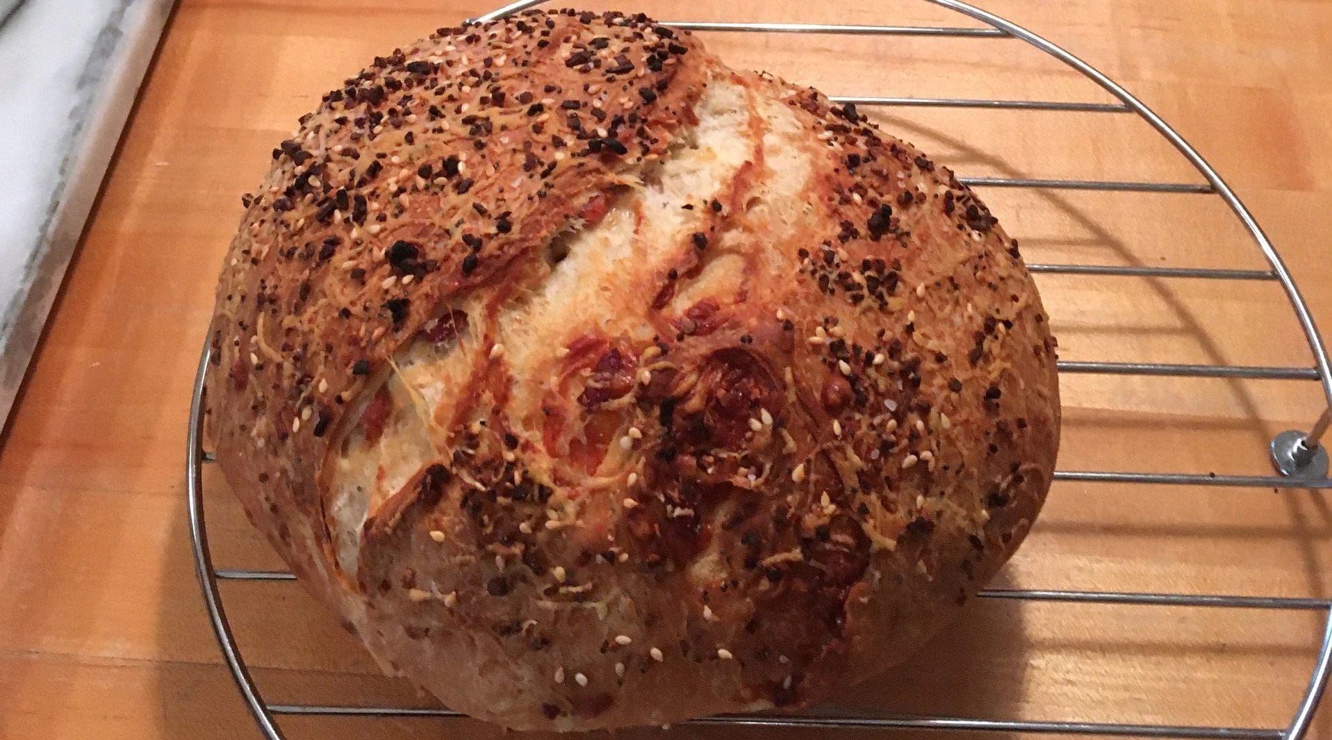 A freshly baked loaf of bread with a crusty top, sprinkled with seeds, resting on a metal cooling rack.