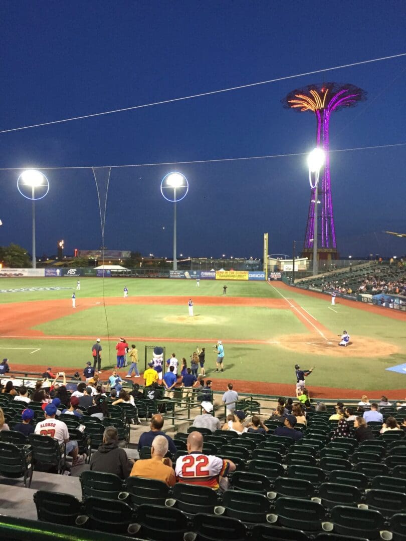 A nighttime baseball game viewed from the stands with a distinctive palm-tree-shaped light structure in the background.