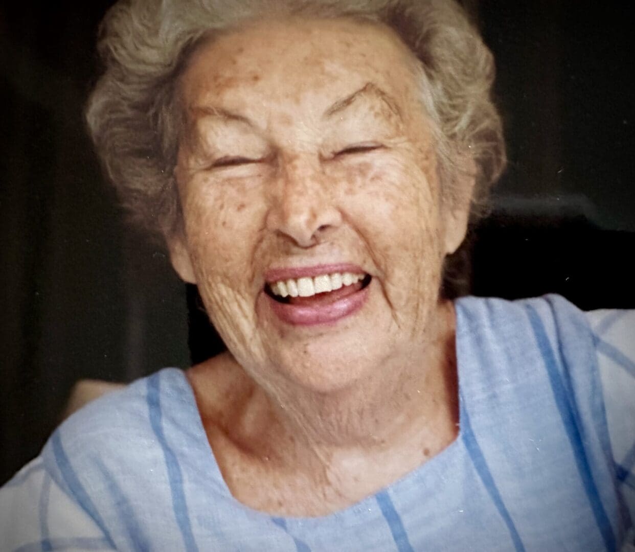 An elderly woman with gray hair, laughing heartily, wearing a blue striped top, viewed through a window.