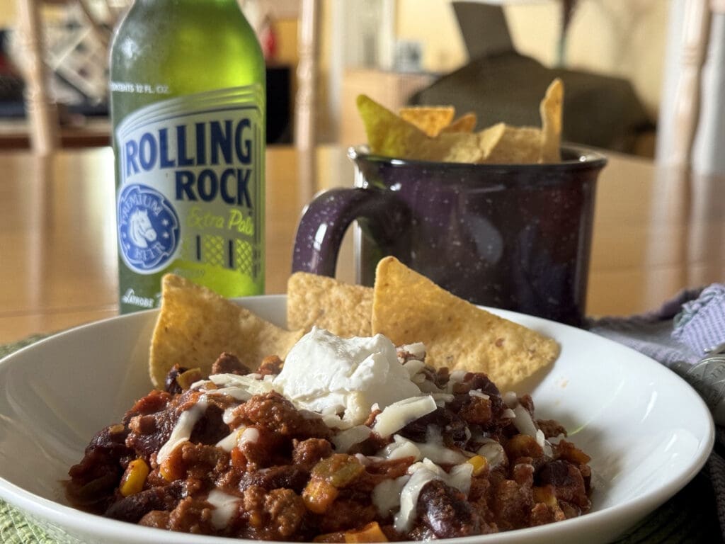 A bowl of chili topped with cheese and sour cream, with tortilla chips and a bottle of rolling rock beer in the background.