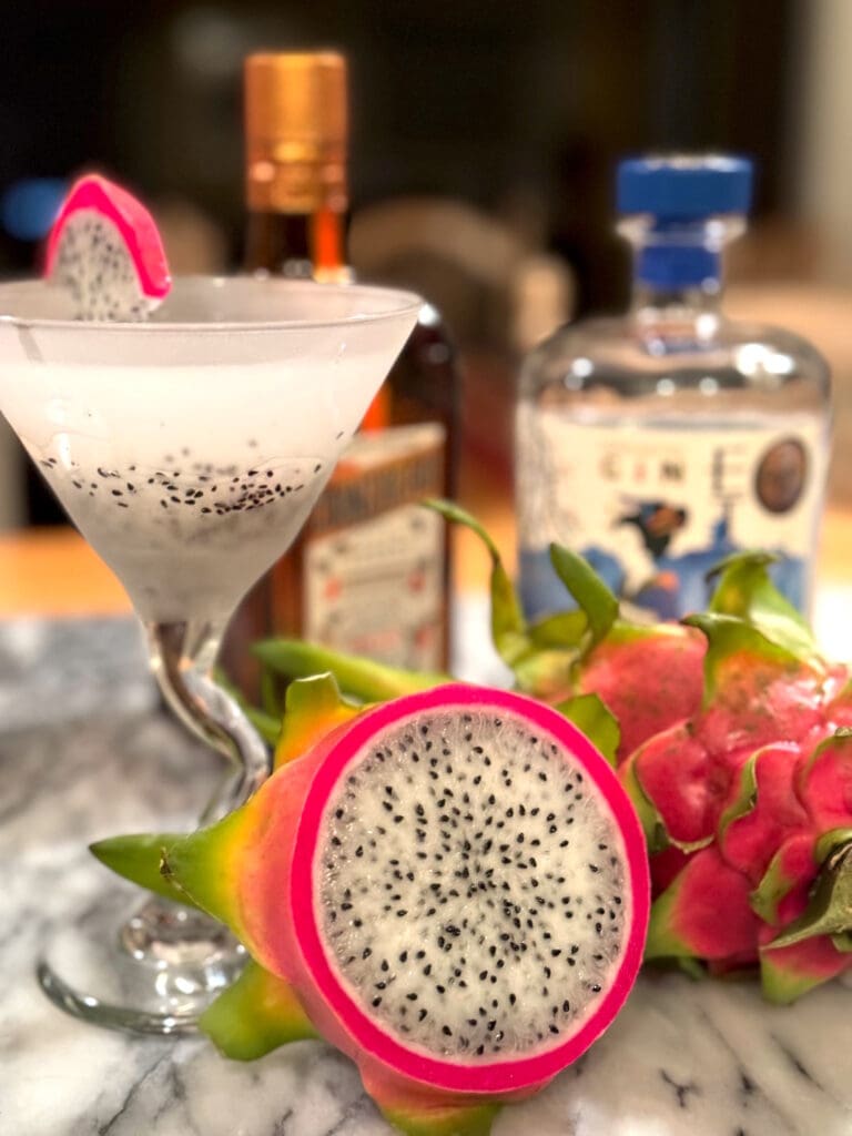 A dragon fruit cocktail garnished with a dragon fruit slice, surrounded by dragon fruits and alcohol bottles on a marble counter.