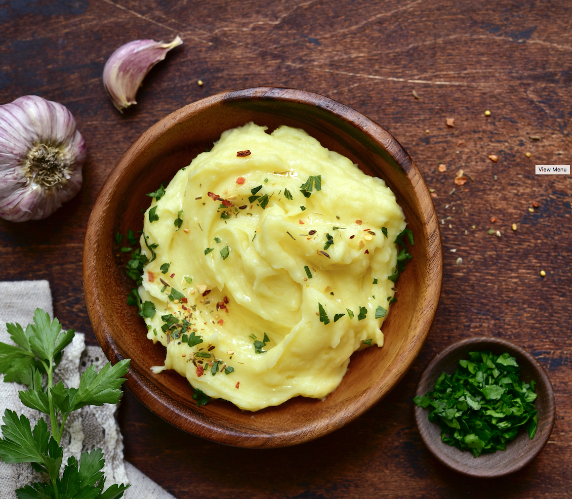 A bowl of creamy mashed potatoes garnished with herbs, served on a wooden table with garlic and parsley nearby.