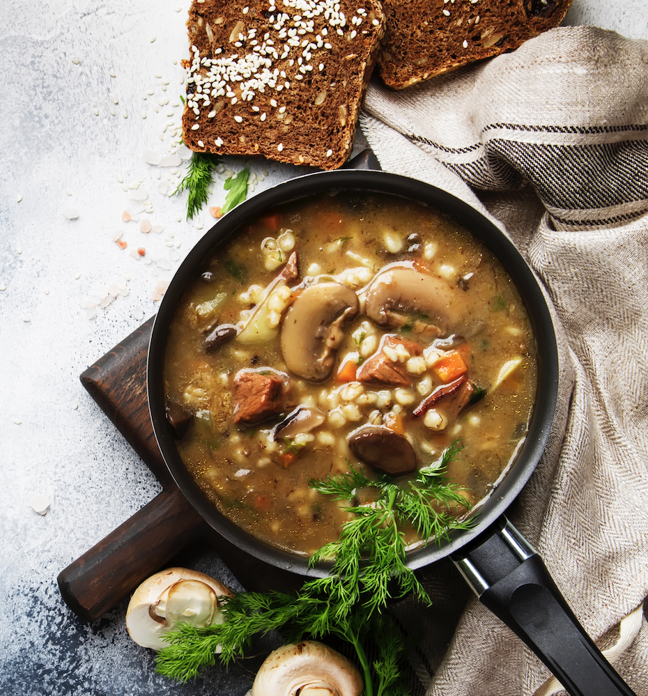 A bowl of mushroom soup garnished with herbs, surrounded by slices of brown bread and fresh mushrooms on a textured surface.