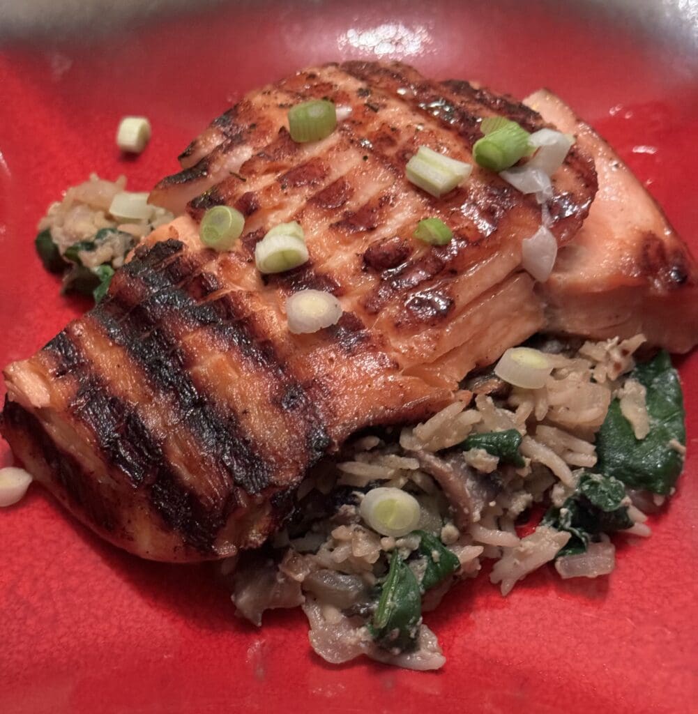 Grilled salmon fillet on a bed of rice with spinach and chopped green onions, served in a red bowl.
