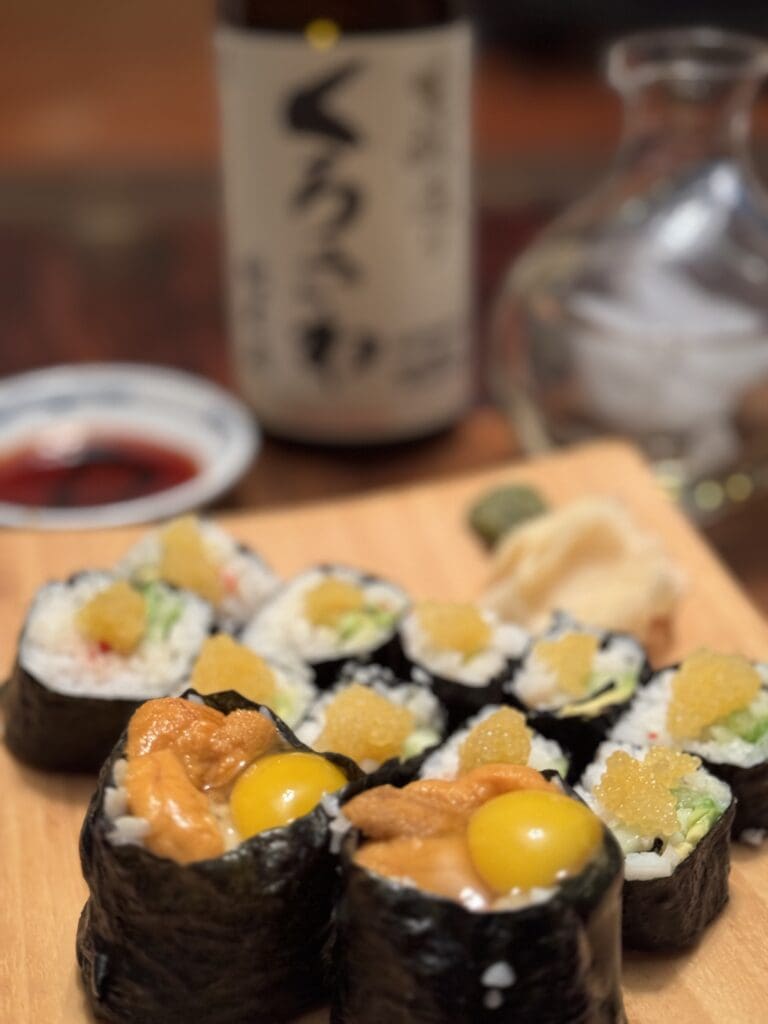 A close-up of sushi rolls garnished with roe and quail eggs, with soy sauce, wasabi, a sake bottle, and a sake decanter blurred in the background.