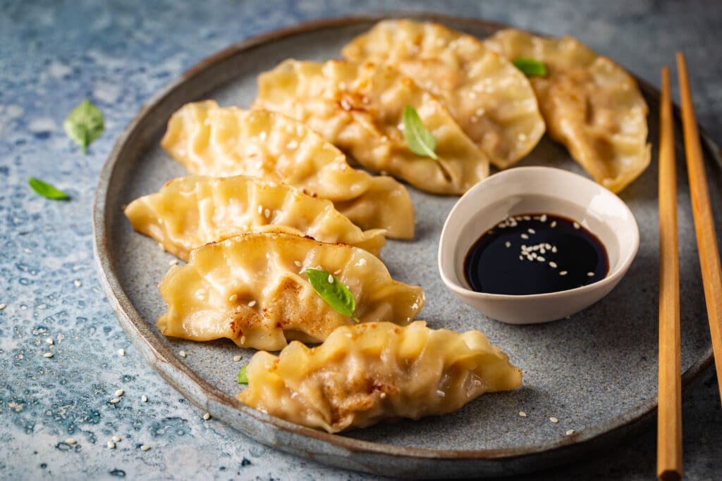 Plate of fried dumplings with a side of soy sauce, garnished with green leaves, on a textured blue surface.