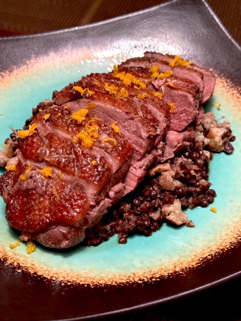 Sliced duck breast with crispy skin served on a bed of black rice, garnished with orange zest on a turquoise plate.