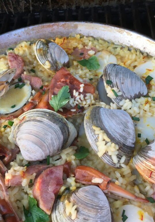 Paella with clams, shrimp, and sausage cooked in a large pan on a grill.