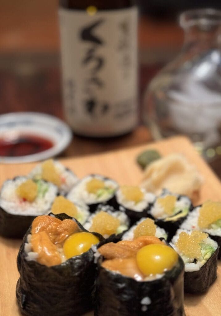 A close-up of sushi rolls garnished with roe and quail eggs, with soy sauce, wasabi, a sake bottle, and a sake decanter blurred in the background.