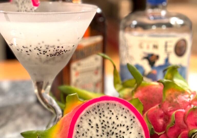 A dragon fruit cocktail garnished with a dragon fruit slice, surrounded by dragon fruits and alcohol bottles on a marble counter.