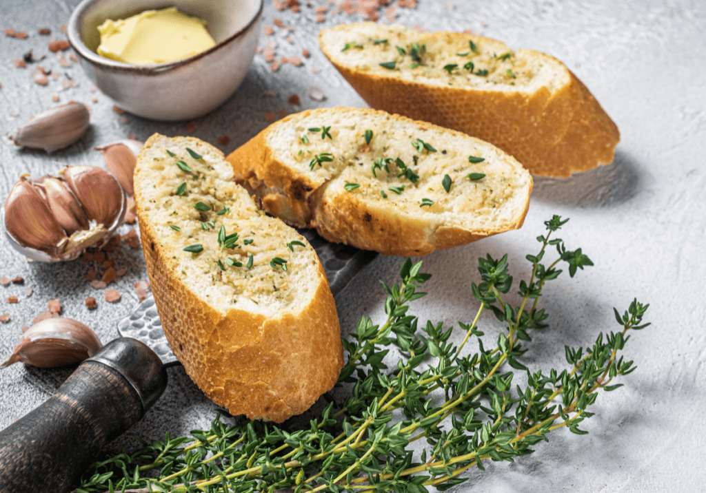 Slices of garlic bread with herbs, butter, garlic cloves, and hazelnuts on a textured surface.