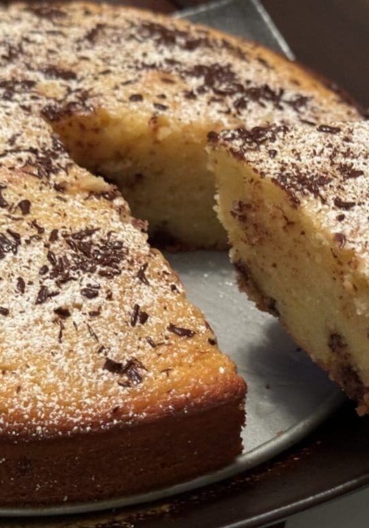 Freshly baked cake on a plate with a slice cut out, sprinkled with powdered sugar and chocolate shavings, under warm lighting.