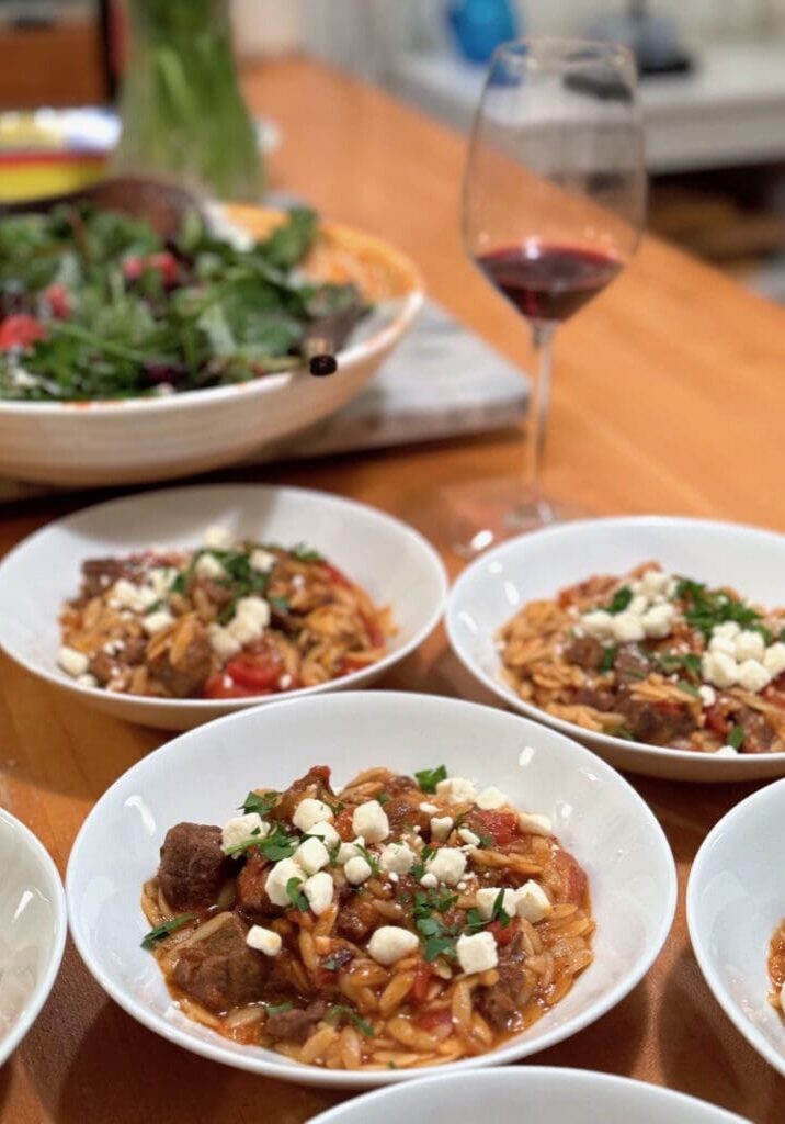 Four plates of pasta with beef and feta cheese, served on a wooden table with a salad bowl and a glass of red wine in the background.