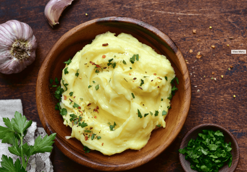 A bowl of creamy mashed potatoes garnished with herbs, served on a wooden table with garlic and parsley nearby.