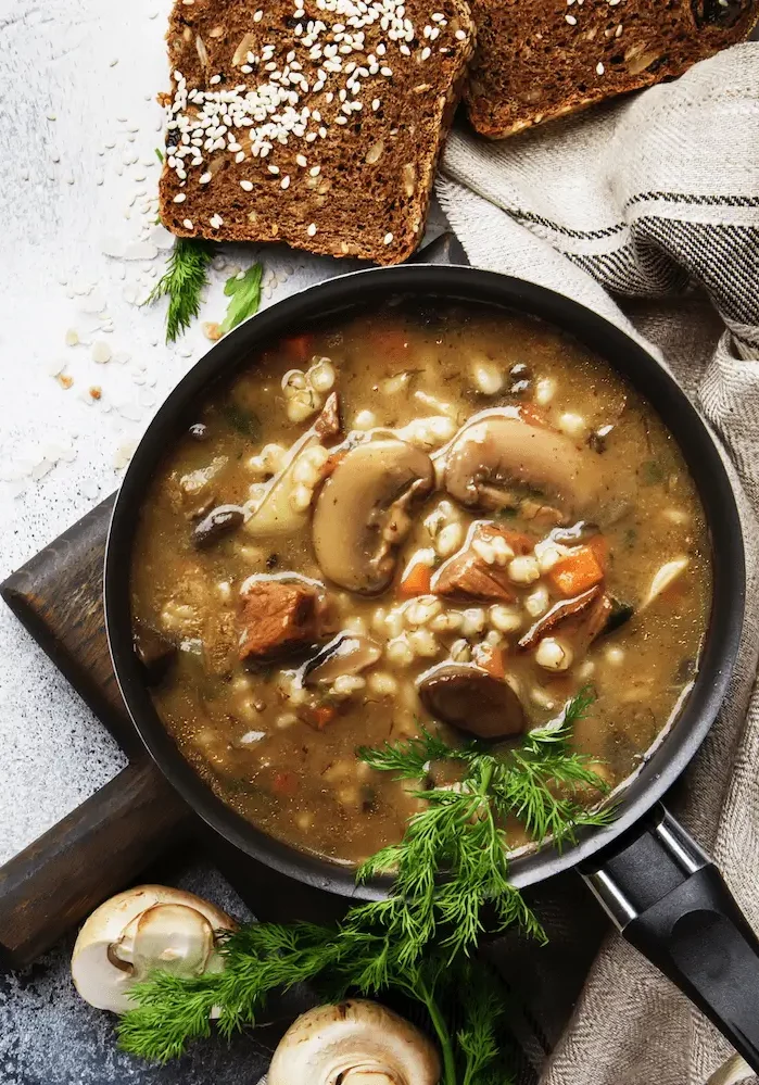 A bowl of mushroom soup garnished with herbs, surrounded by slices of brown bread and fresh mushrooms on a textured surface.