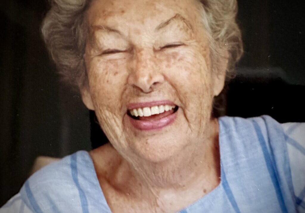 An elderly woman with gray hair, laughing heartily, wearing a blue striped top, viewed through a window.