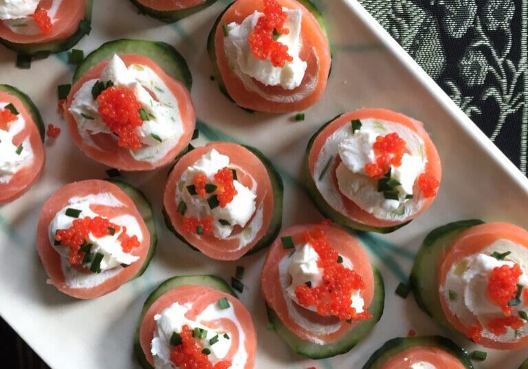 A plate of salmon and cream cheese appetizers topped with caviar and herbs, served on slices of cucumber.