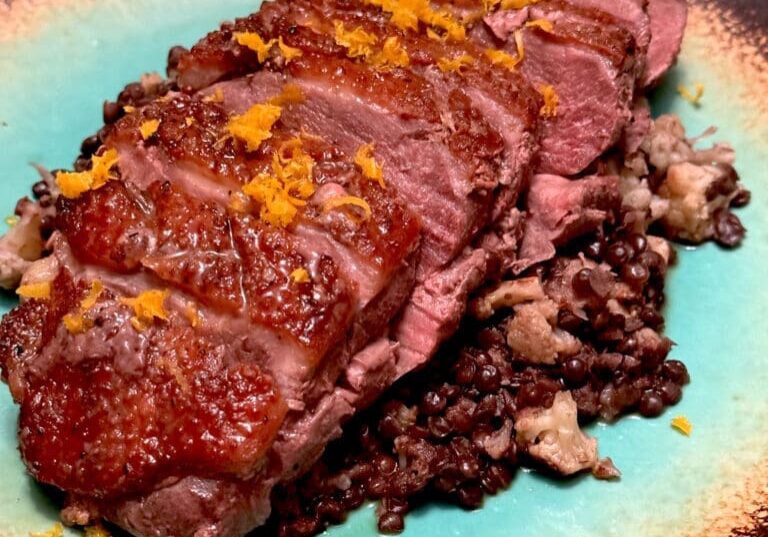 Sliced duck breast with crispy skin served on a bed of black rice, garnished with orange zest on a turquoise plate.